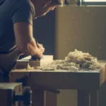 A craftsman skillfully shaping wood at a workshop bench with wood shavings visible.