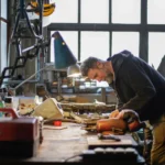 Side view of concentrated adult bearded male mechanic in black workwear standing near workbench in lamplight and fixing metal detail while working in workshop
