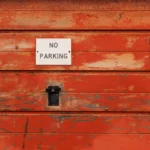 A rustic red wooden door with a no parking sign attached.