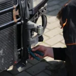 Mechanic working on car repair outdoors using tools, focusing on vehicle radiator area.