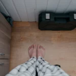 Overhead view of bare feet in pajamas on wooden floor indoors with tool box nearby.