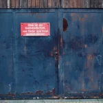 An aged metal garage door with a humorous sign in German about parking.