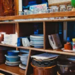 A wooden kitchen shelf stocked with assorted dishes, glasses, and condiments. Cozy vintage style.