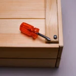 Orange screwdriver on a wooden box corner with screw detail.