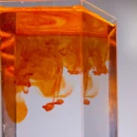 Glass with pure water and diffusing orange paint streams with blots on light background