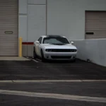 A white Dodge Challenger sports car parked in an urban industrial area with rolled-up garage doors.