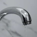 A close-up shot of a modern chrome bathroom faucet with water dripping from the tip.