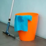 Close-up of cleaning supplies including orange bucket, mop, and blue rag indoors.